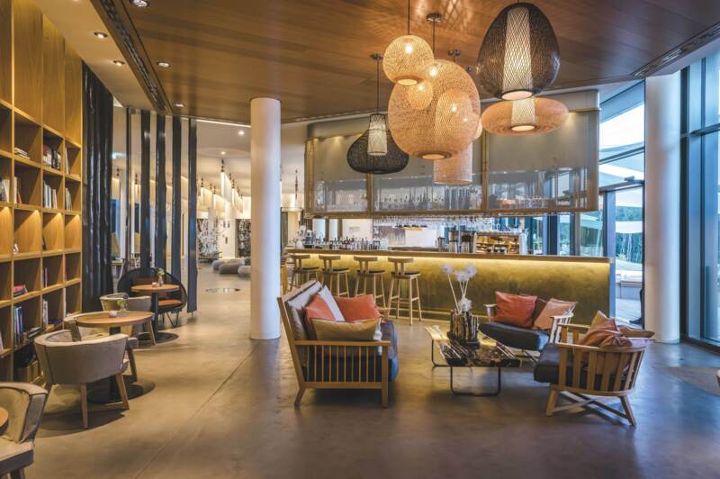 Zicht vanuit de library naar de bar en lobby. Natuurlijke materialen als hout, steen en linnen zorgen voor een warme, uitnodigende sfeer, terwijl verfijnde designelementen het eigentijdse karakter benadrukken 